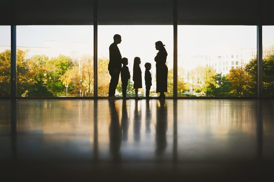 Silhouettes of a family with two children stand facing each other in front of a large window, with a park visible outside.