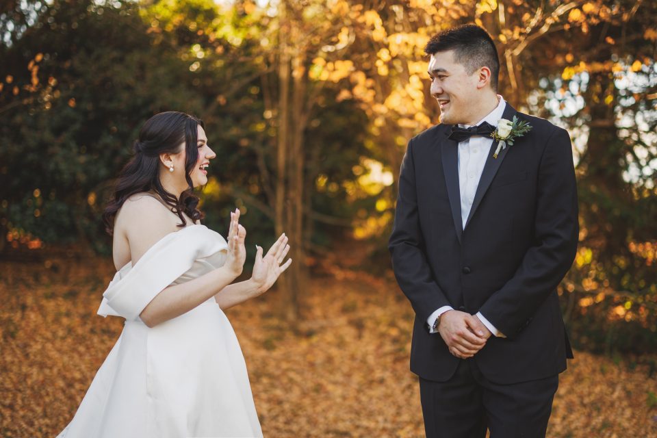 A bride and groom smile at each other outdoors, standing on fallen leaves with trees in the background. The bride gestures with her hands while the groom stands with a boutonniere on his tuxedo.