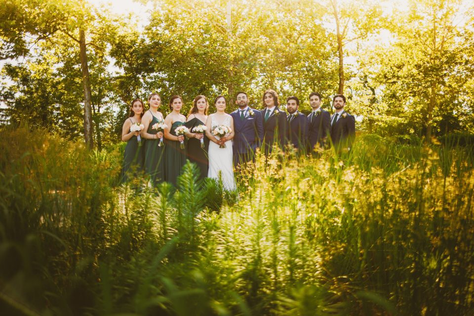A bridal party stands outdoors among greenery. The bride is in a white dress, surrounded by bridesmaids in dark green dresses and groomsmen in dark suits.