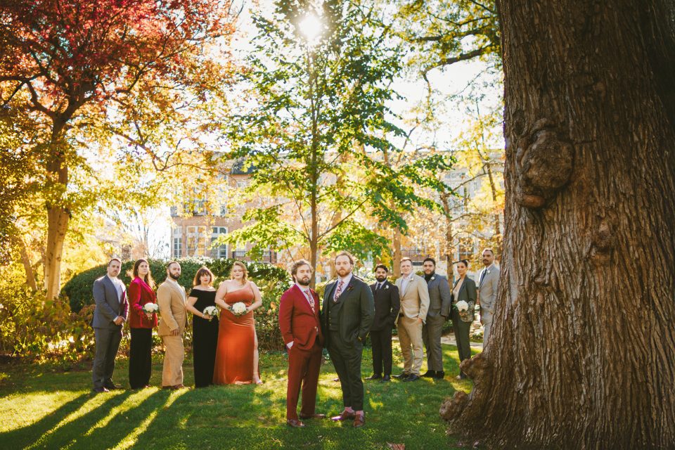A group of people dressed formally stand in a line outdoors, surrounded by trees with autumn leaves.