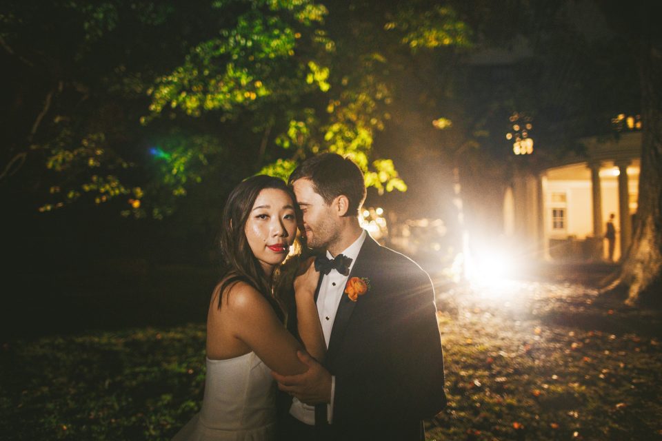 A bride and groom embrace outdoors at night, dressed in formal attire, with illuminated foliage and a building in the background.
