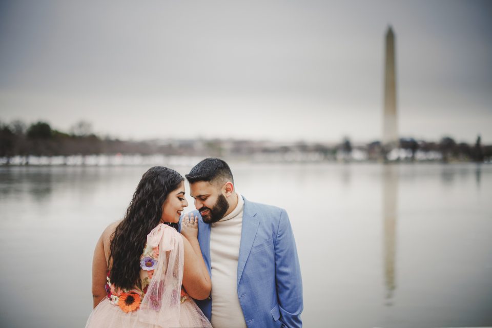 A couple stands by a reflective body of water, with a tall monument visible in the background. The woman wears a floral dress, and the man is in a blue blazer.