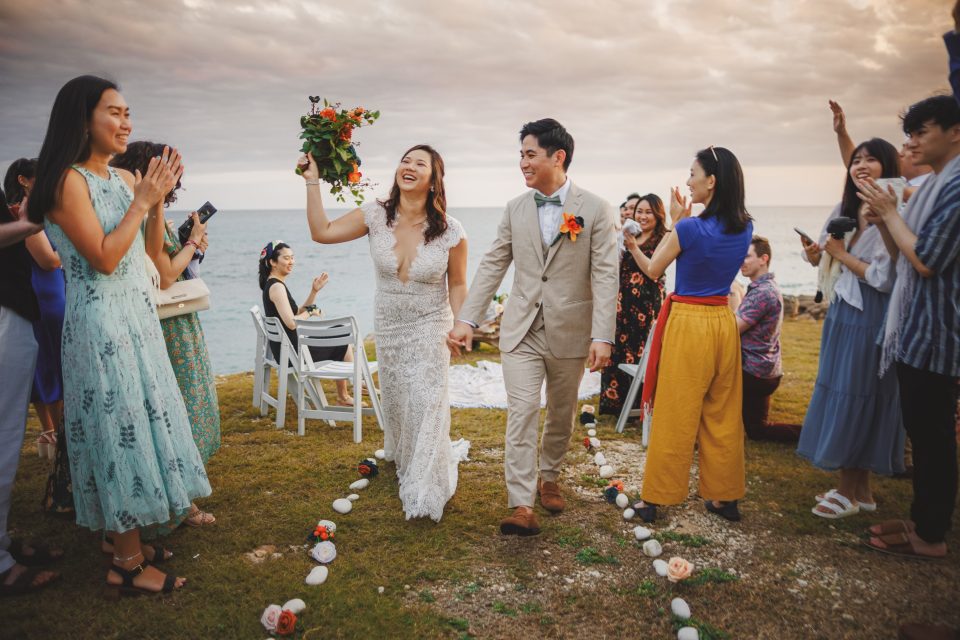 A couple walks hand-in-hand down a makeshift aisle outdoors, surrounded by clapping friends. The woman holds a bouquet. The setting is near the sea under a cloudy sky.
