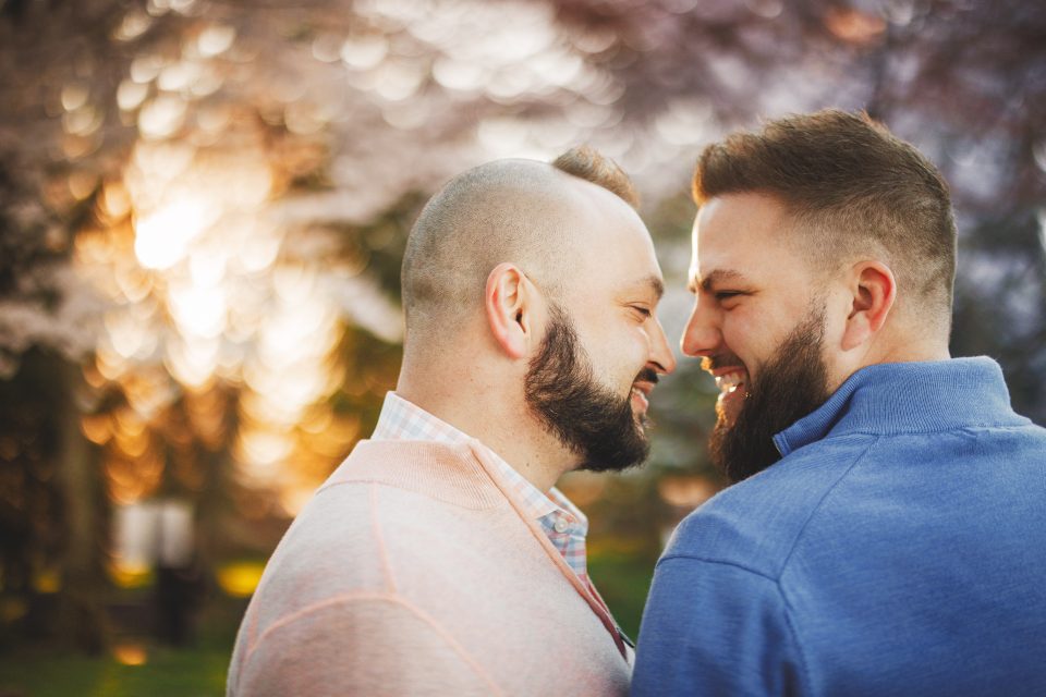 Two men stand closely, smiling at each other outdoors, with a blurred background of sunlight and trees.