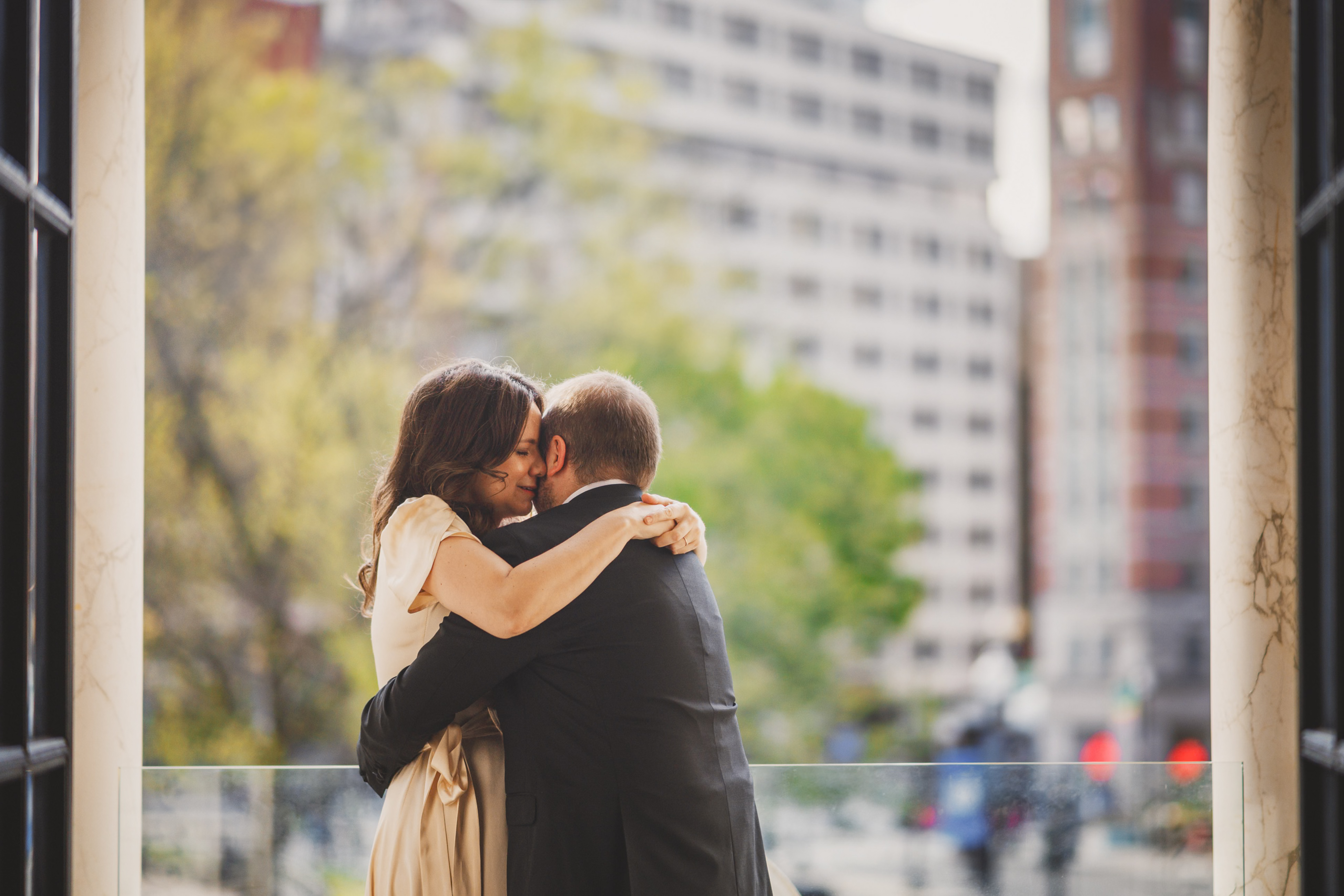A couple embraces on a balcony overlooking a cityscape with blurred buildings and trees in the background.