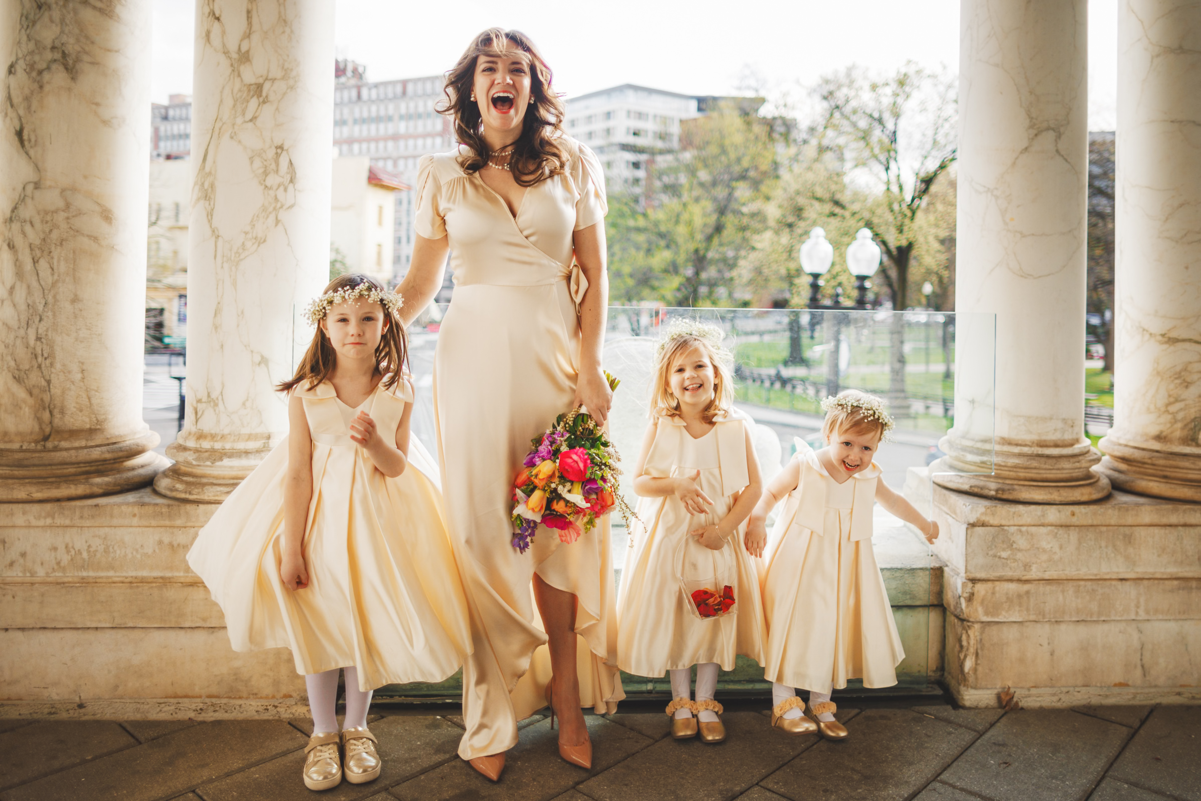 A woman in a cream dress stands with three young girls in matching dresses, each holding bouquets, on a balcony with columns.