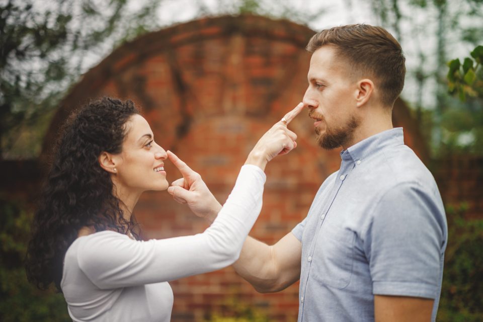 Two people standing outside, facing each other with smiles, touching each other's noses with their index fingers. A brick wall is in the background.