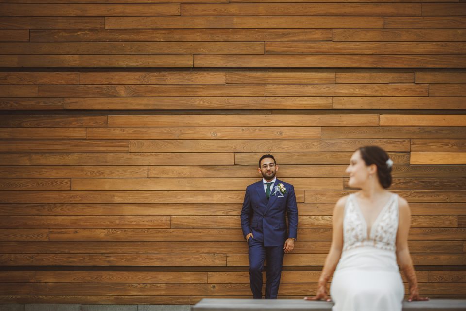 A bride sits on a bench in the foreground, wearing a white dress. A groom in a blue suit stands against a wooden wall in the background.