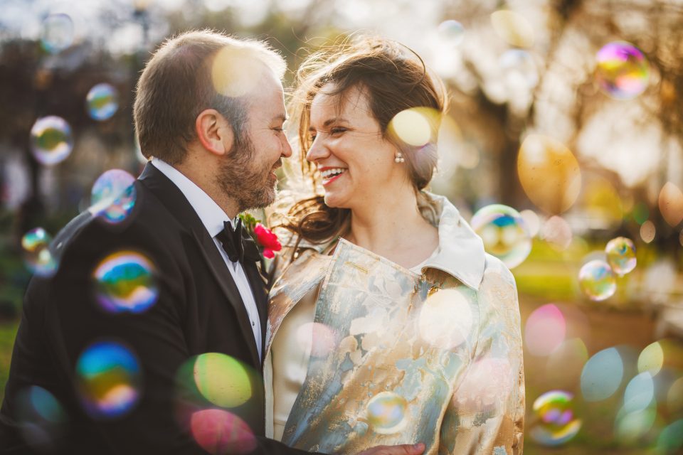 A couple smiles at each other surrounded by bubbles outdoors. The man is in a suit, the woman in a light-colored jacket. It's a sunny day.