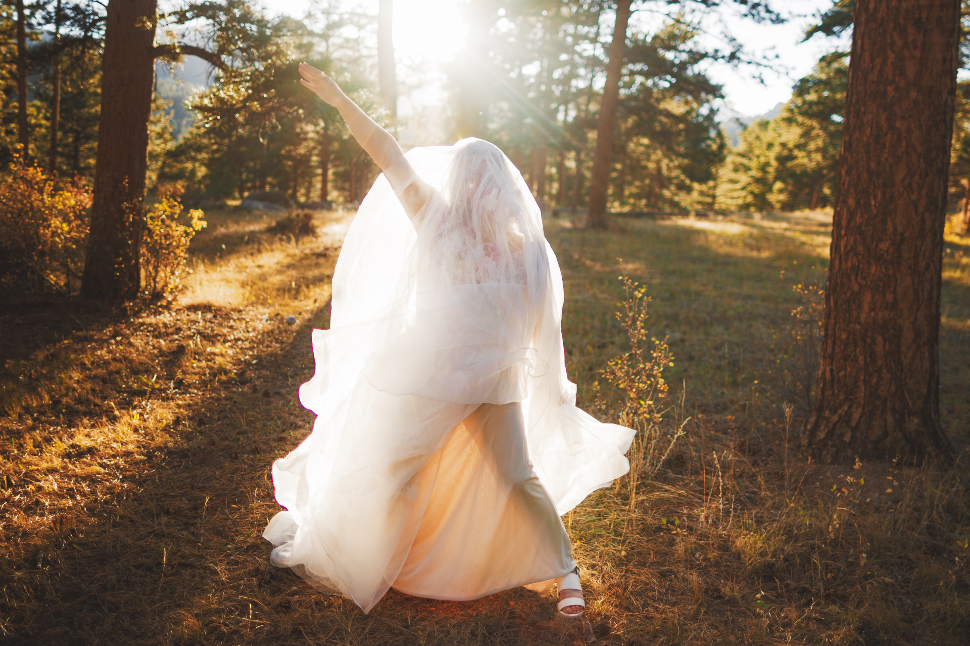 Bride in a white dress and veil poses energetically in a sunlit forest clearing, surrounded by trees.