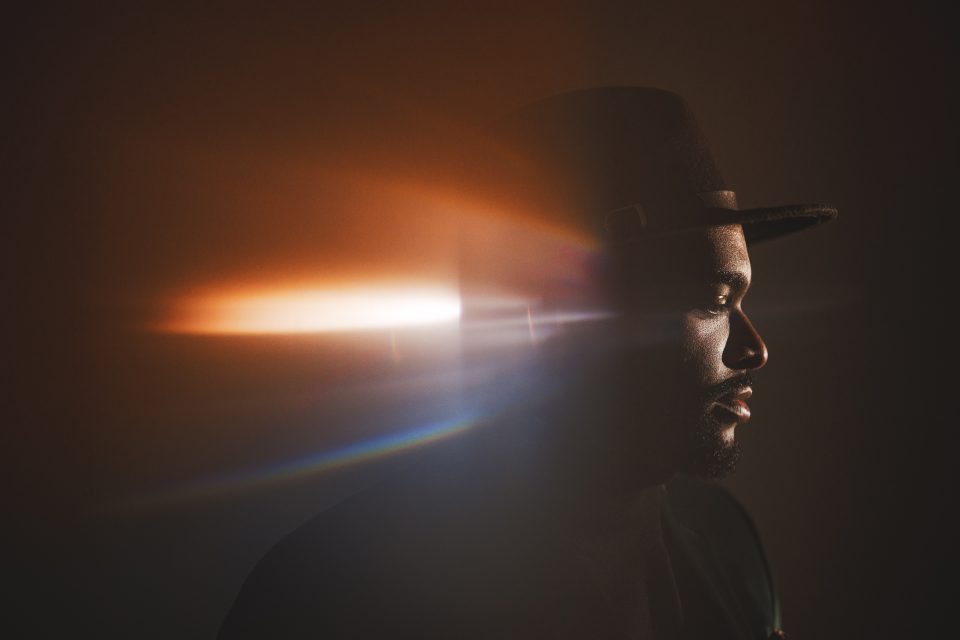 Silhouetted man wearing a hat, with dramatic lighting creating a lens flare effect across the image.