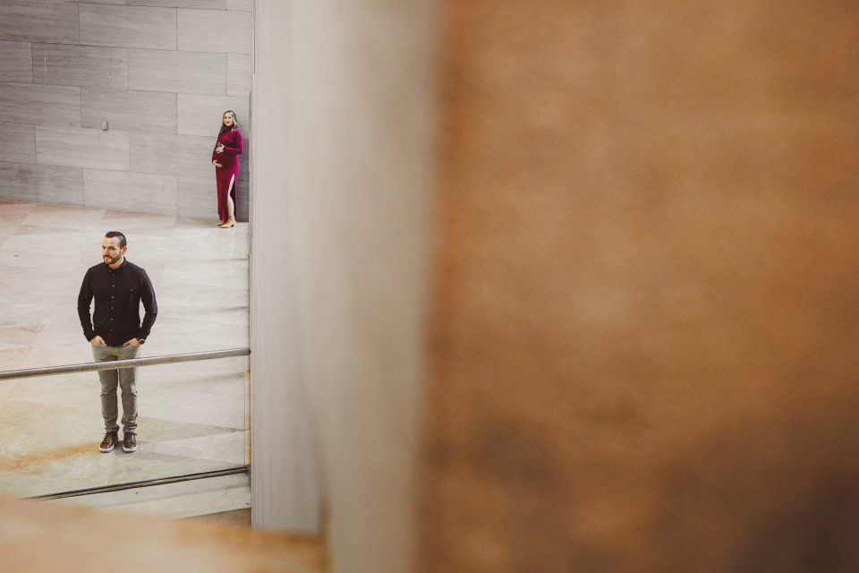 Two people stand apart in a spacious, modern interior with concrete walls. The man is in the foreground, wearing casual clothes, while the woman in the background leans against a wall.