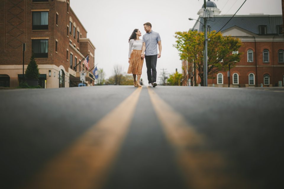 A man and woman walk hand in hand down an empty street, with buildings and autumn trees in the background.