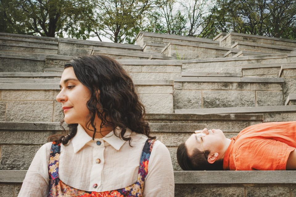 Woman sitting on stone steps in profile with another person lying down in an orange shirt, looking upward. Trees are in the background.