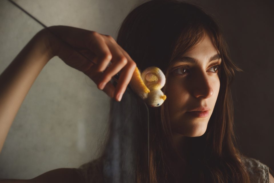 A woman holds a small, yellow knitted mouse by a string, hanging it in front of her face. The lighting is dim and moody.