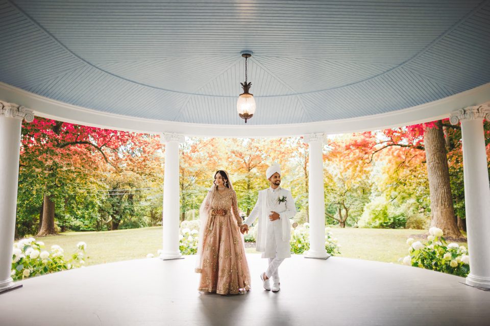 A couple in traditional attire stands under a gazebo, surrounded by autumn trees.