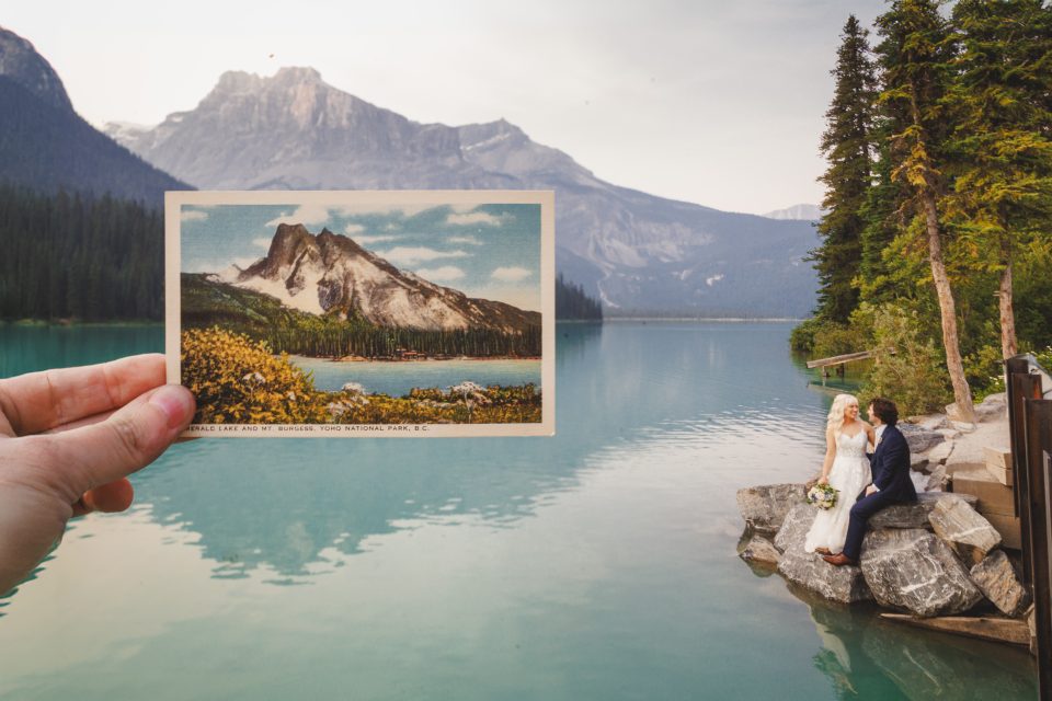 Hand holding a vintage postcard of a mountain and lake, aligned with the actual scenery. A couple in wedding attire sits on rocks by the lake.