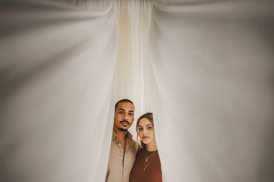A man and woman stand closely together, framed by parted white curtains, looking directly at the camera.