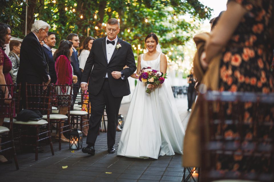 A bride in a white dress holds a bouquet and walks down an aisle lined with seated guests, accompanied by a man in a tuxedo. The setting is outdoors with string lights and greenery.