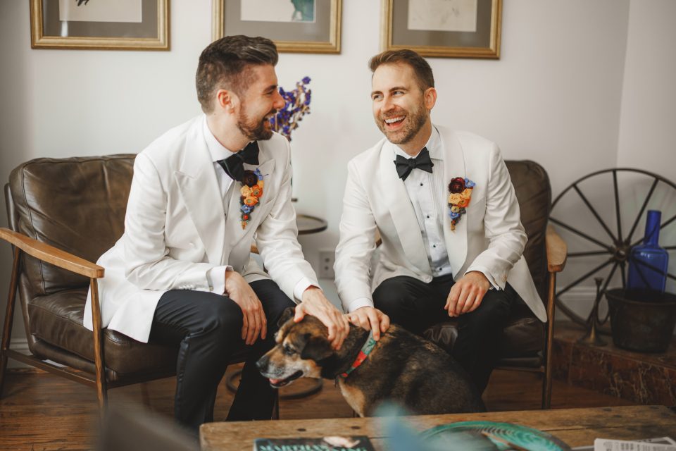 Two men in white tuxedos sit on a sofa, smiling and petting a dog. They wear flower boutonnieres and are in a room with wall art and a decorative wheel.