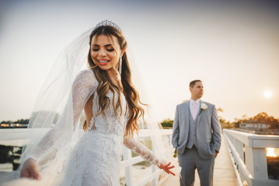 Bride in a white gown with a veil stands on a bridge, smiling with eyes closed. A groom in a gray suit stands behind her, looking to the side. Sunset in the background.