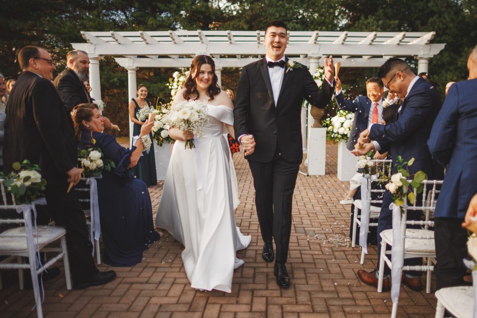 Bride and groom smile and hold hands, walking down the aisle outdoors, surrounded by seated guests.