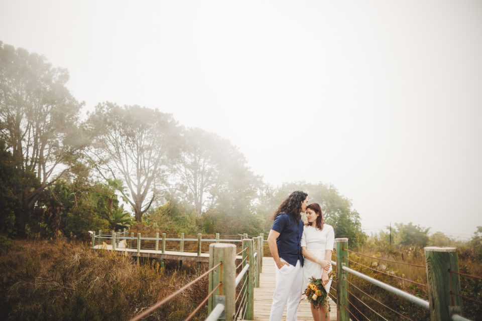 Couple standing on a wooden boardwalk in a foggy park, surrounded by trees. The woman holds a bouquet of flowers.