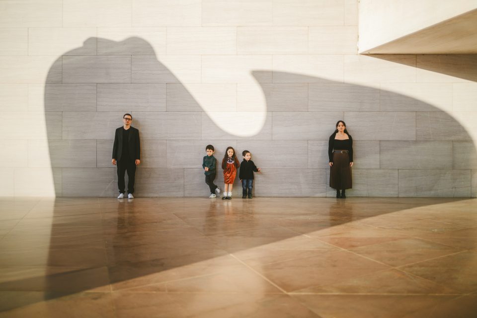 Two adults and three children stand against a stone wall. The shadow of a large sculpture creates a dramatic effect over the scene.