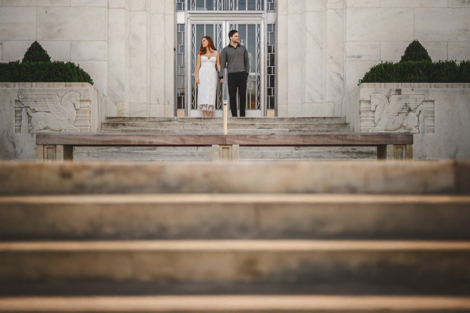 A man and woman stand at the top of stone steps in front of a large building with a metal door and sculptures.