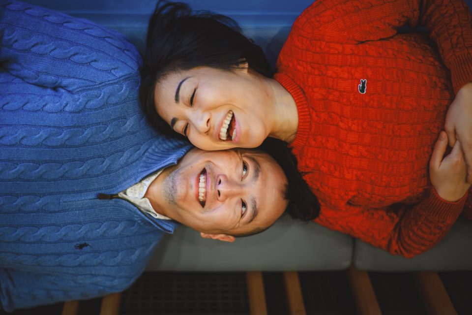 A man and a woman, both wearing knit sweaters, lie on a wooden bench, smiling and looking in different directions.