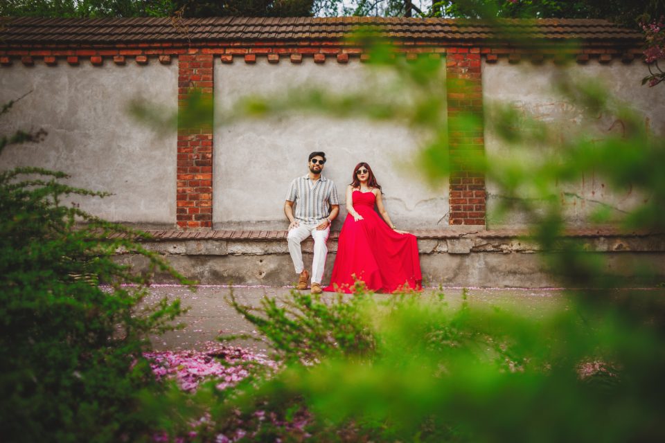 A man and a woman sit on a bench beside a brick and plaster wall, surrounded by green foliage and pink flowers. The woman wears a red dress; the man wears a striped shirt and white pants.