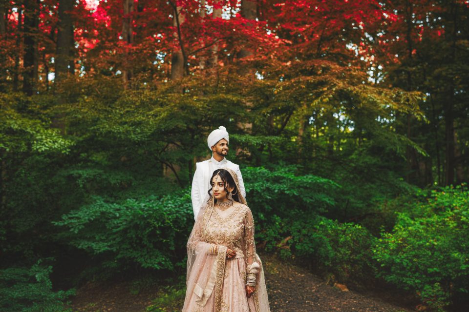 A couple stands in a lush forest with vibrant red and green foliage. The man wears a white suit and turban, while the woman is in a detailed pink and gold dress with a matching scarf.