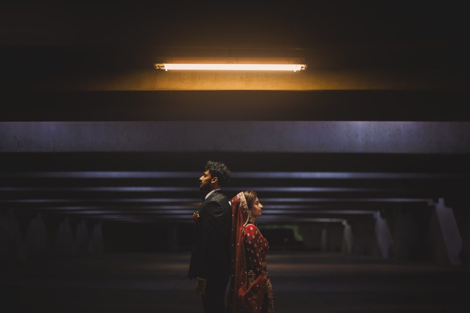 A man and a woman stand back-to-back under a bright fluorescent light in a dimly lit space.
