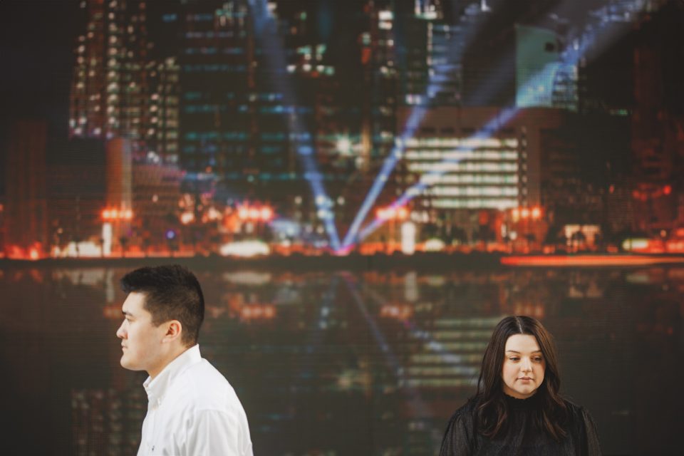 Two people stand apart in front of a cityscape background with bright lights and reflections.