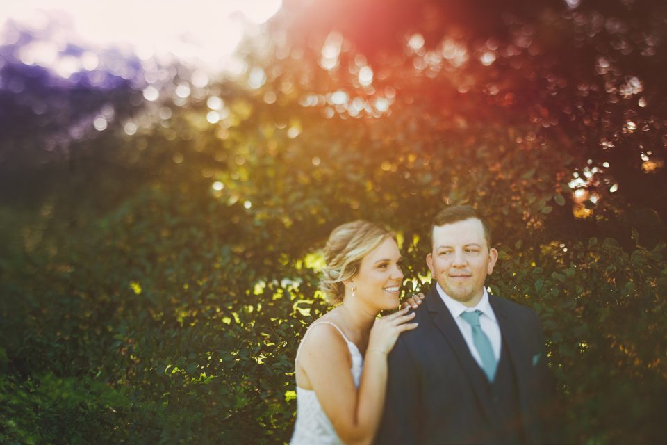 A bride and groom stand together in a garden setting, with sunlight filtering through the trees in the background.