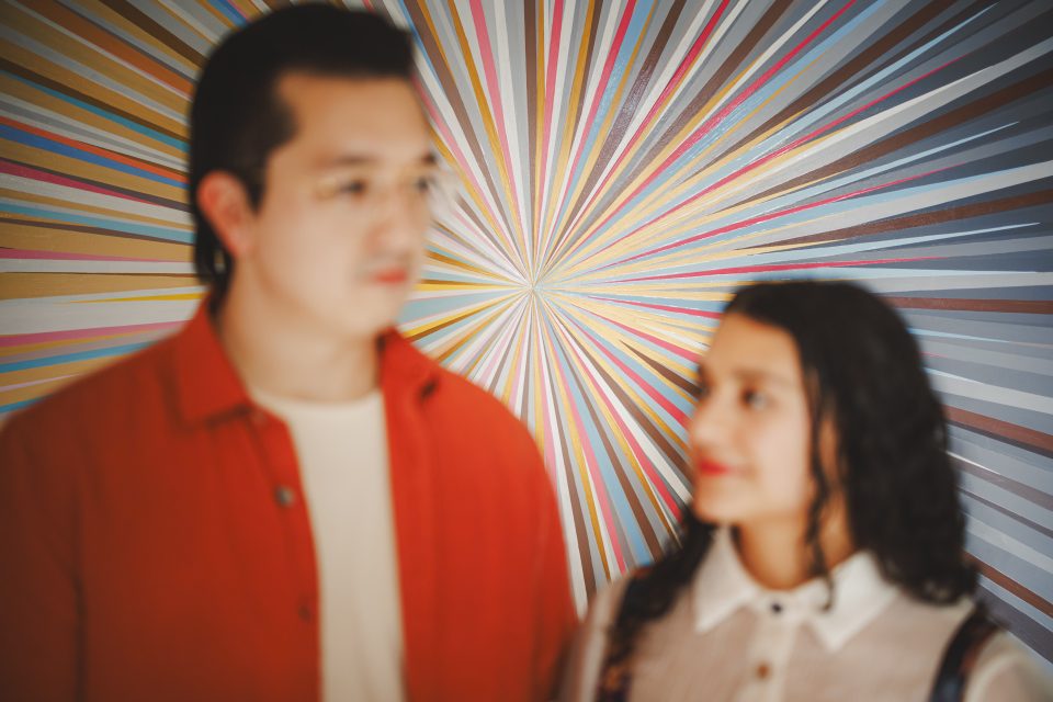 A man and woman stand in front of a colorful radial striped background, slightly out of focus, facing each other.