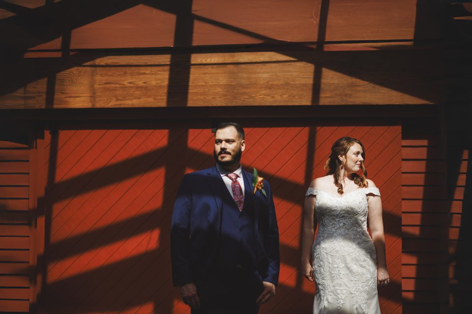 A couple stands side by side, dressed in wedding attire. The groom is in a dark suit, and the bride is in a white gown. They are under diagonal shadows against a red wooden background.