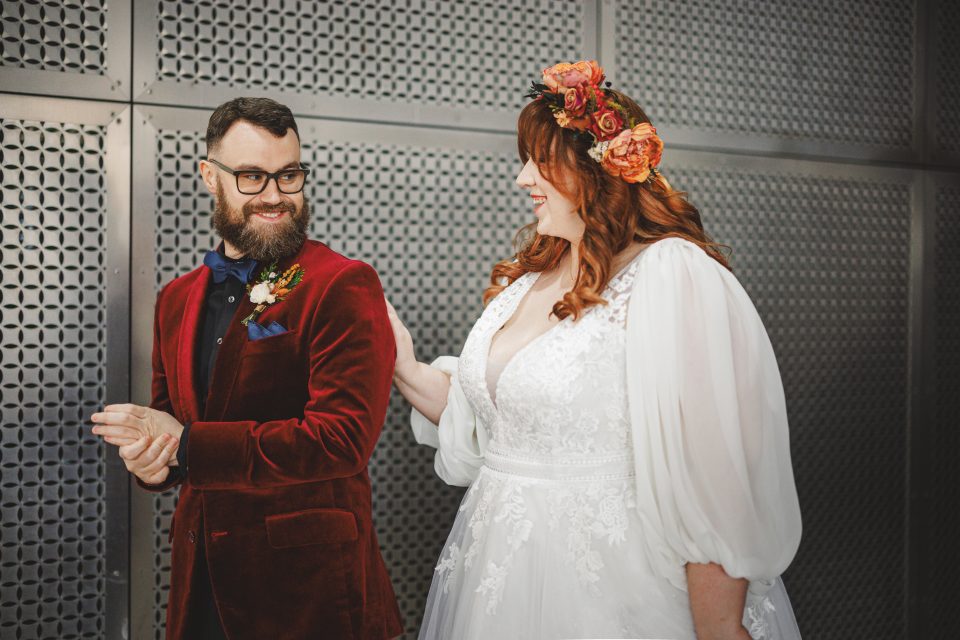 A couple standing together; the man wears a red velvet suit and the woman wears a white dress with floral headpiece.
