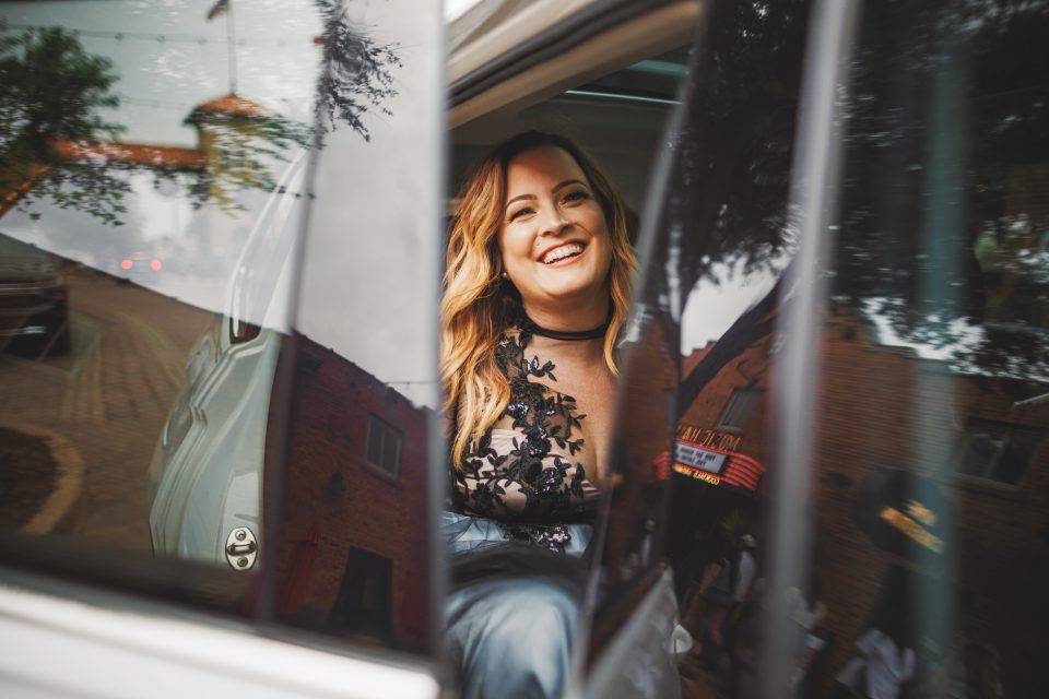 Woman smiling while seated in a car, partially outside, with the car door open. Reflections of trees and buildings visible on the car windows.