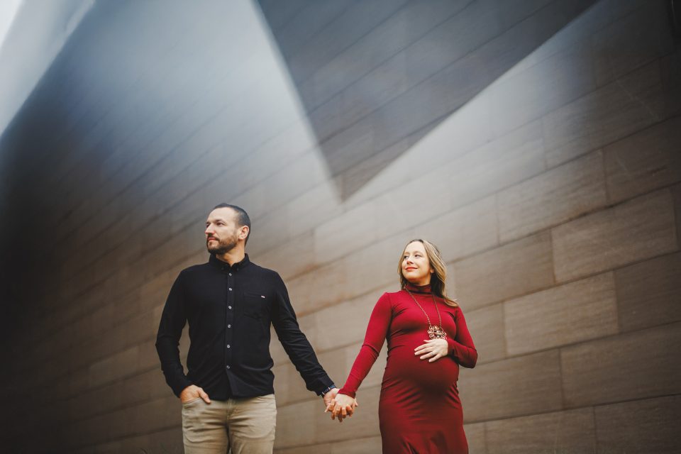 A pregnant woman in a red dress holds hands with a man in a black shirt. They walk along a modern, sunlit corridor.
