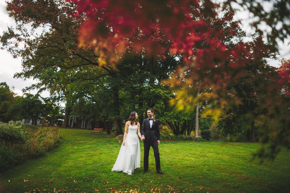 A bride and groom hold hands while standing on a grassy lawn, surrounded by trees with autumn leaves.