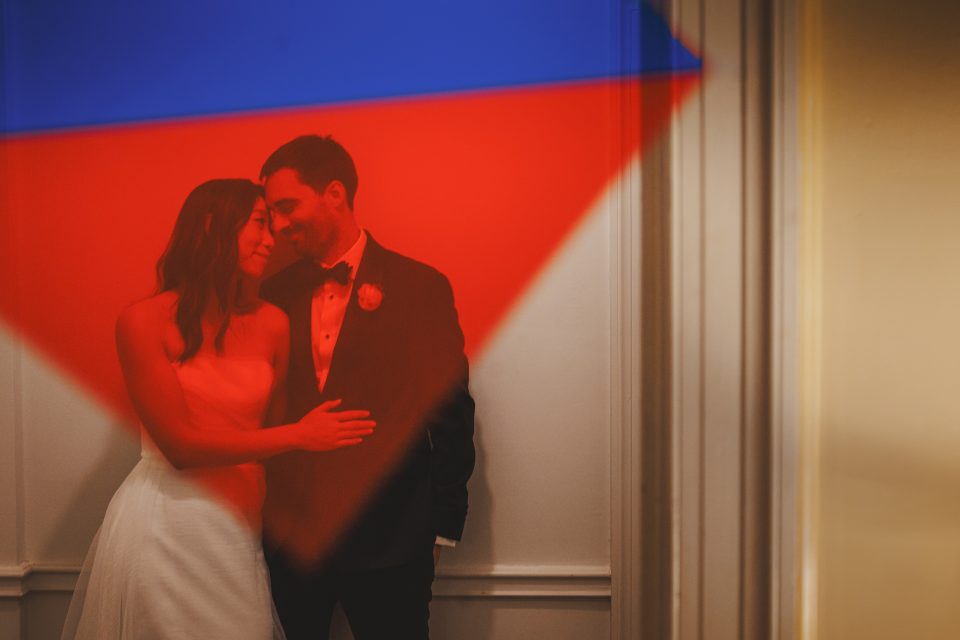 Bride and groom stand closely under a red and blue light projection, casting colorful shadows on the wall behind them.