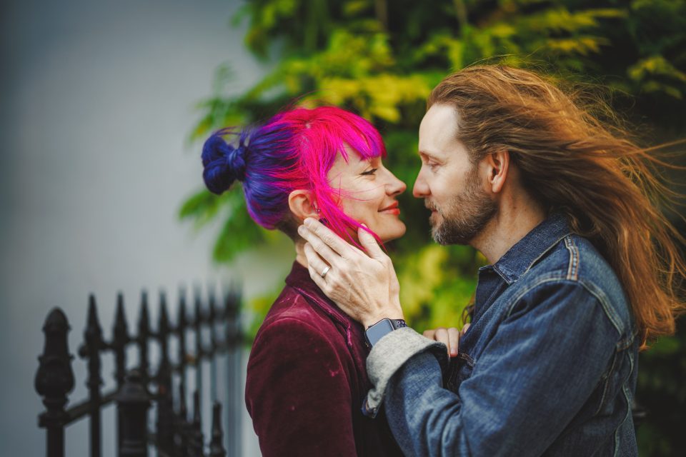 A couple with colorful hair faces each other closely, smiling. The woman has vibrant pink and purple hair. The man has long brown hair. A wrought iron fence and greenery are visible in the background.