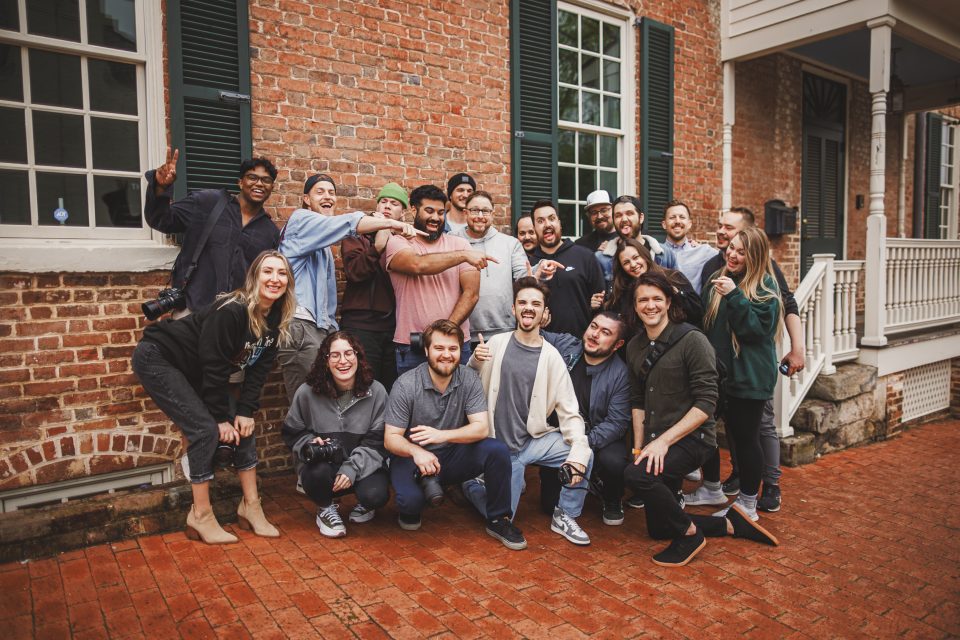 A group of twenty people posing together in front of a brick building with green shutters and a porch. They are smiling and making various hand gestures.
