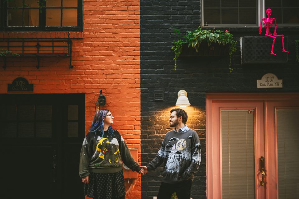 A couple stands holding hands outside. The building behind them is orange and black with a potted plant and a small pink skeleton decoration on an air conditioning unit above.