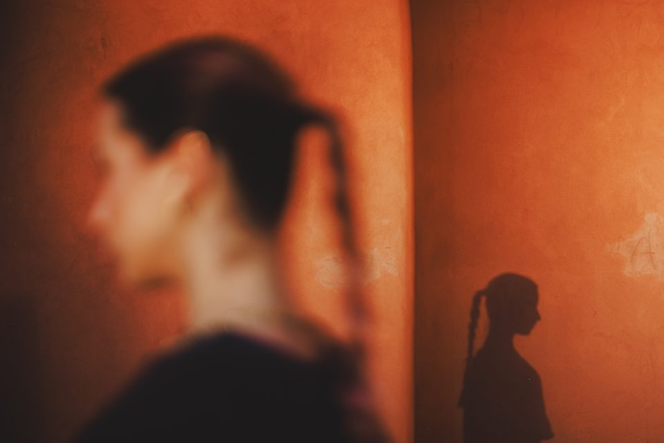A person with a ponytail is seen in profile against an orange wall, with their shadow cast clearly on the wall.
