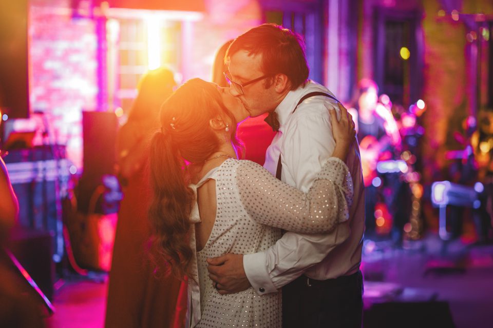 A couple kisses on the dance floor at an event, surrounded by colorful lights and a live band in the background.