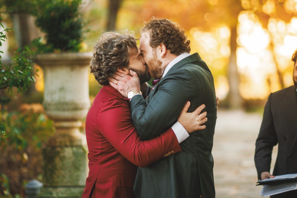 Two men in suits kissing outdoors, surrounded by autumn foliage.