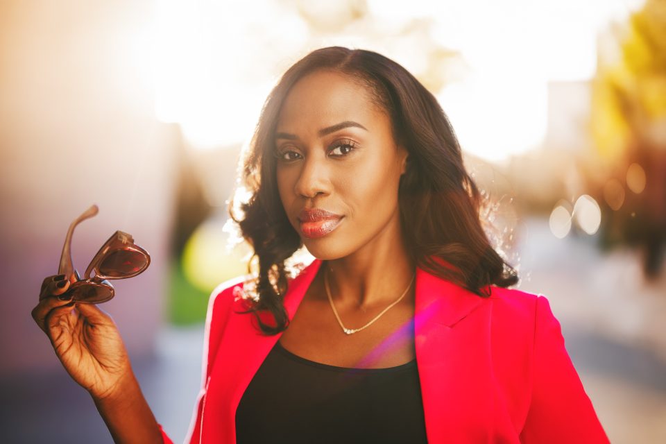 Person outdoors, wearing a red jacket, holding sunglasses in one hand, with a blurred background and sunlight.