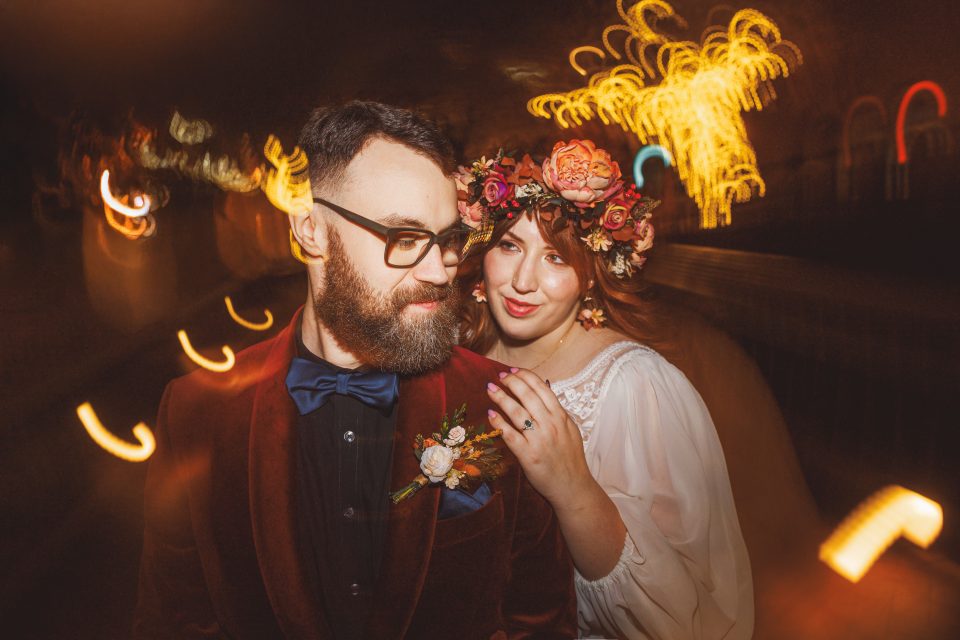 A couple poses at night. The man wears glasses and a velvet suit with a bow tie. The woman, in a white dress, wears a floral crown. Blurred lights create a warm, dreamy background.